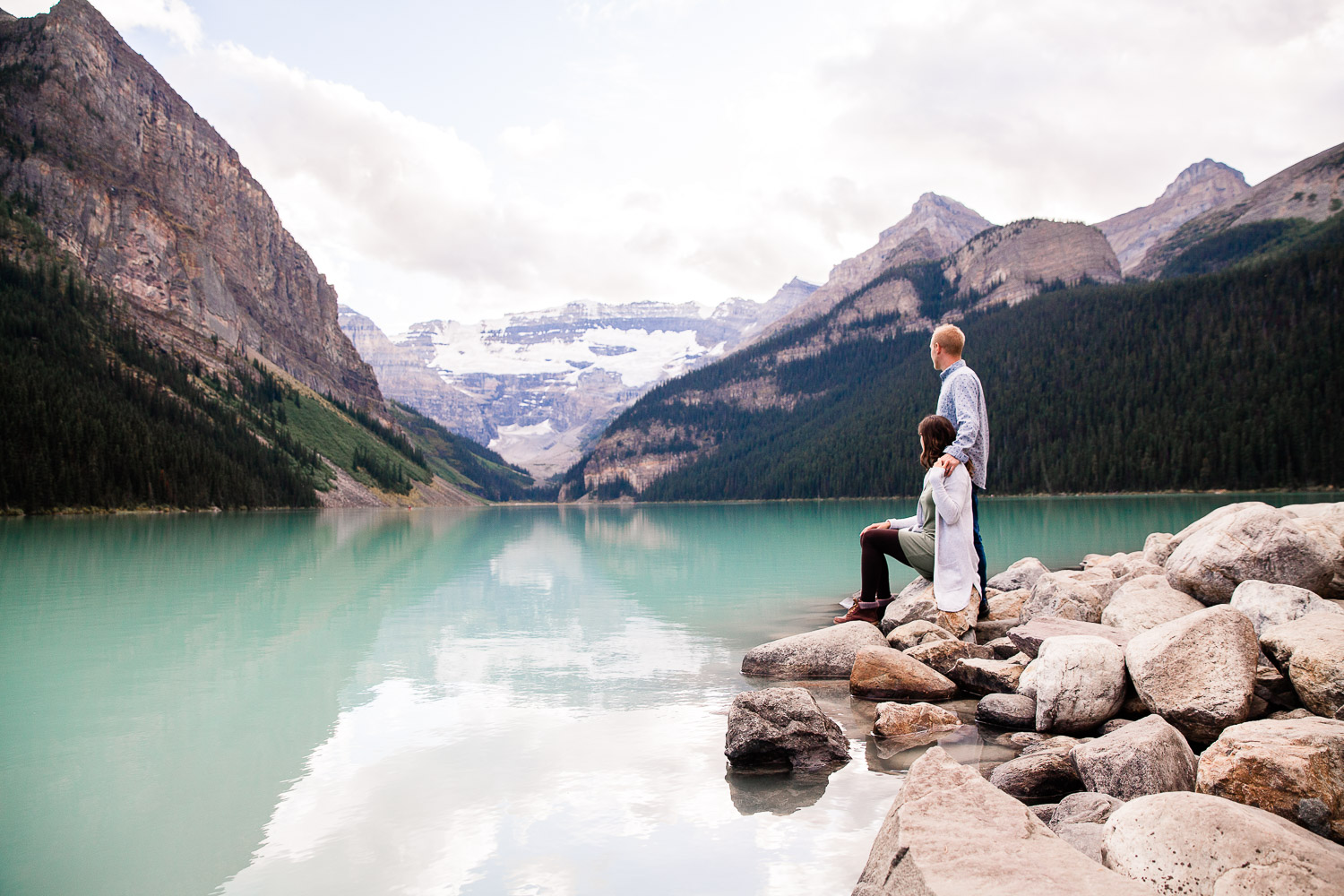 couple standing on rocks while looking at lake louise with mountains in background
