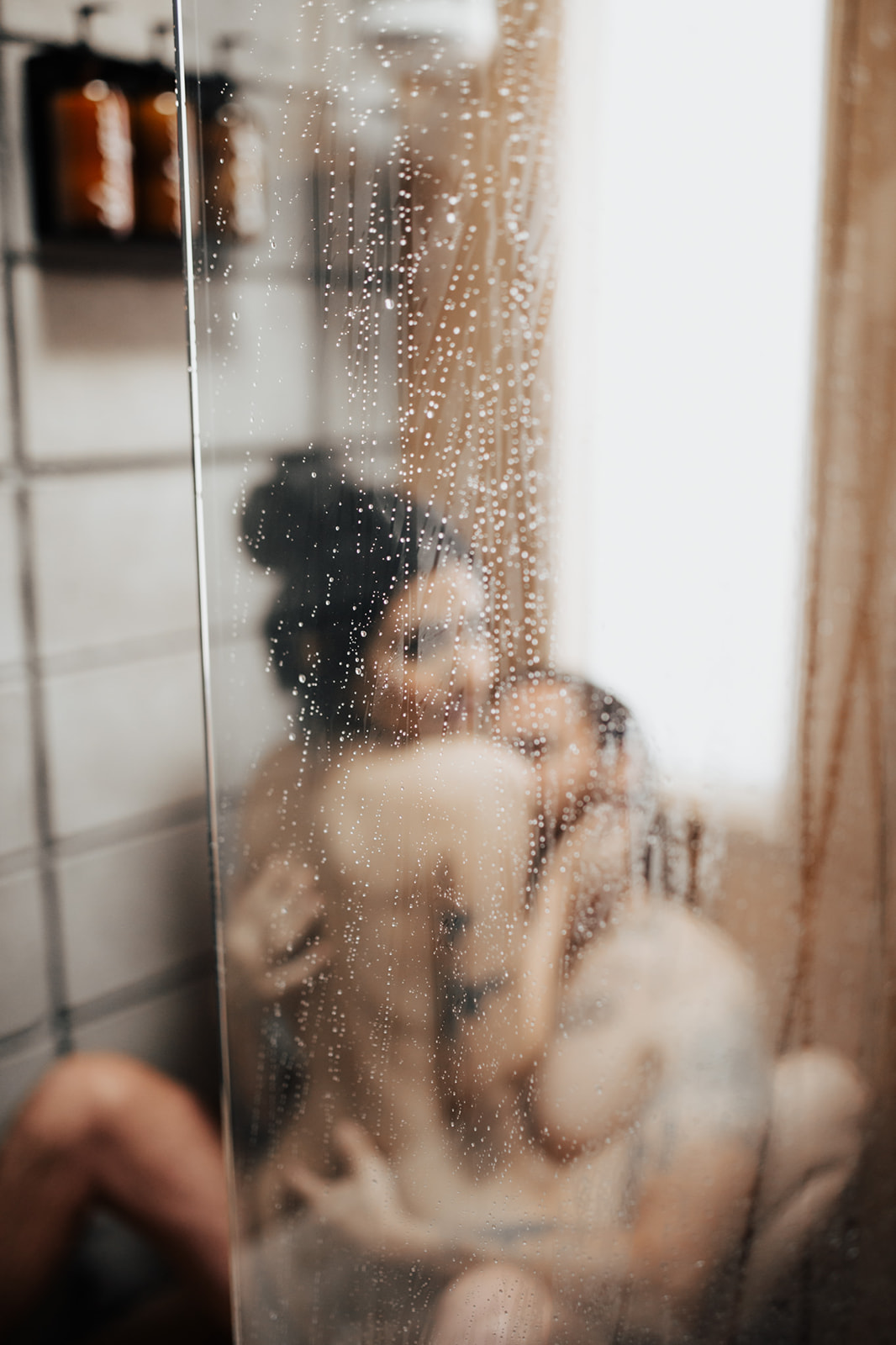 Woman sitting in mans lap while in shower hidden by blurry glass wall with water drops