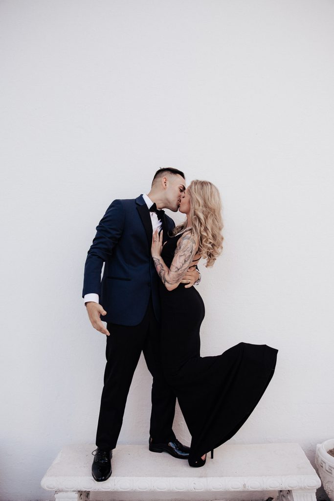 Very chic couple in blue tuxedo and black dress standing on bench in front of white wall an kiss.