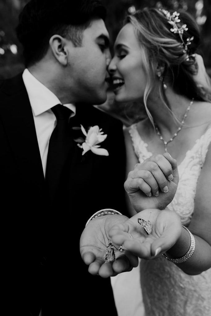 Black and white photo. Bride and groom kiss while holding butterflies in their hands at Magnolia Plantation
