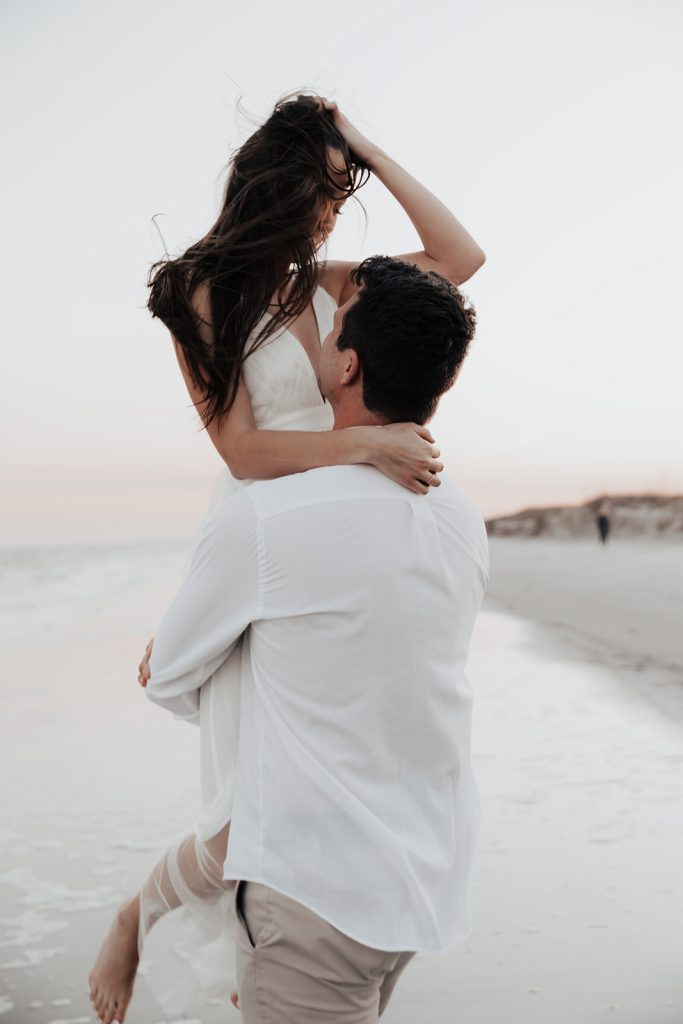 Man lifts up fiancé in white dress at beach photo session