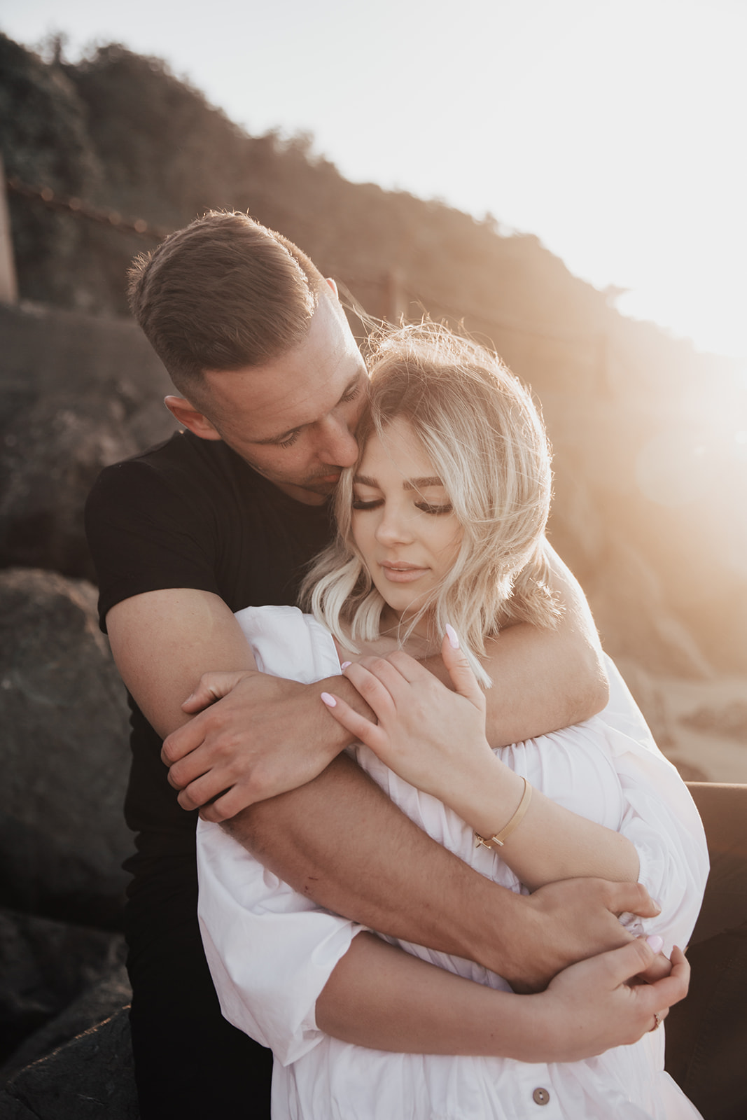 dreamy golden gate engagement. Man wraps arms around woman in white dress with sunset in background