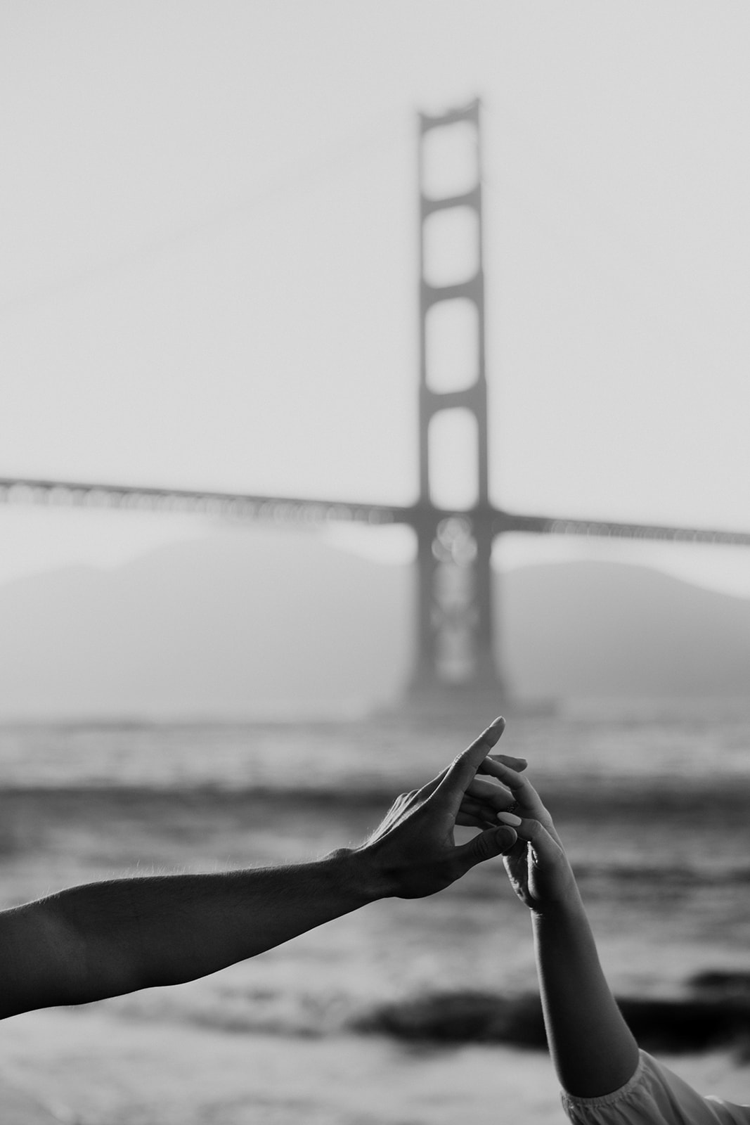 black & white photo of his and hers hand touching with golden gate bridge in background