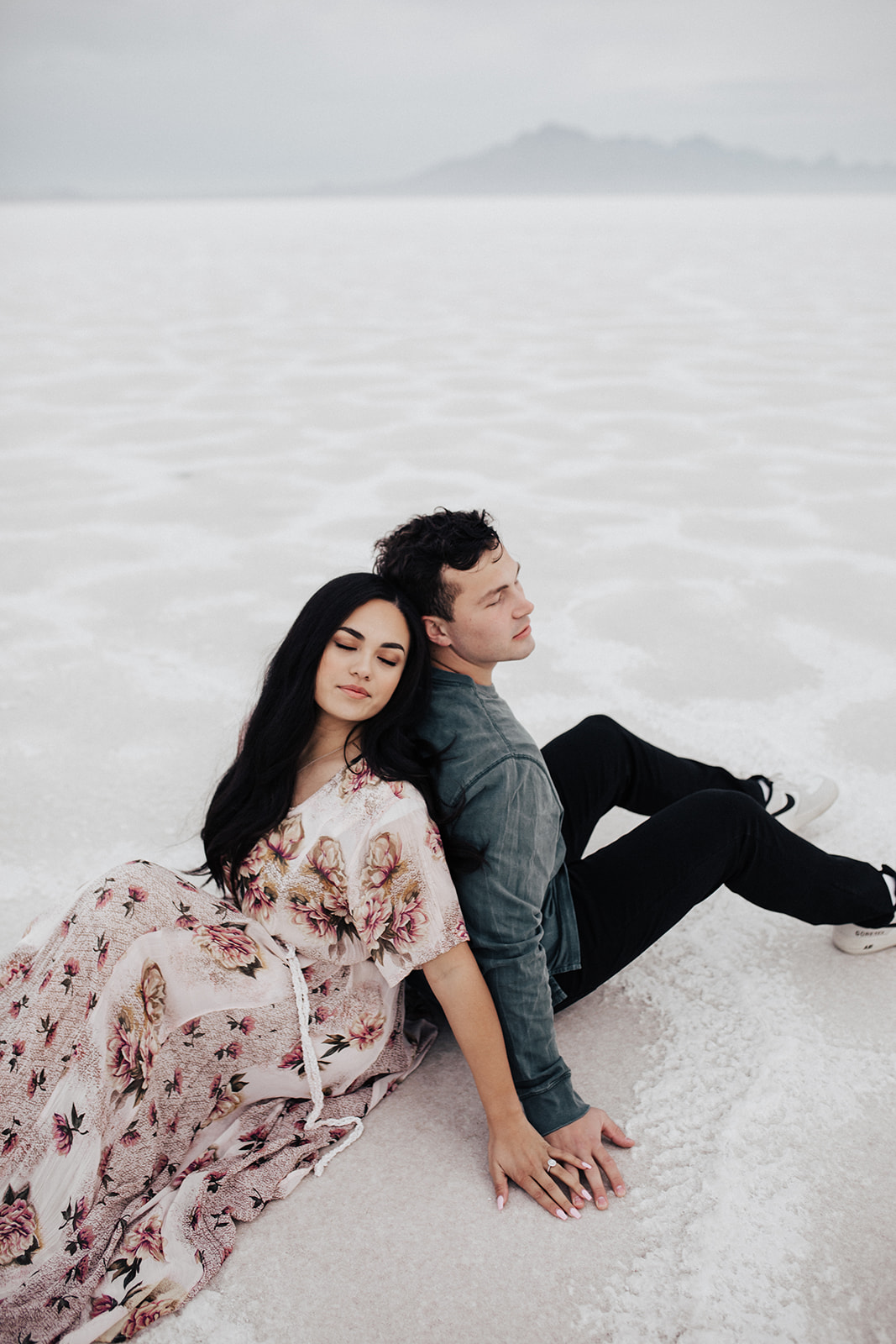 Couple sitting back to back at Bonneville Salt Flats with eyes closed for engagement photos