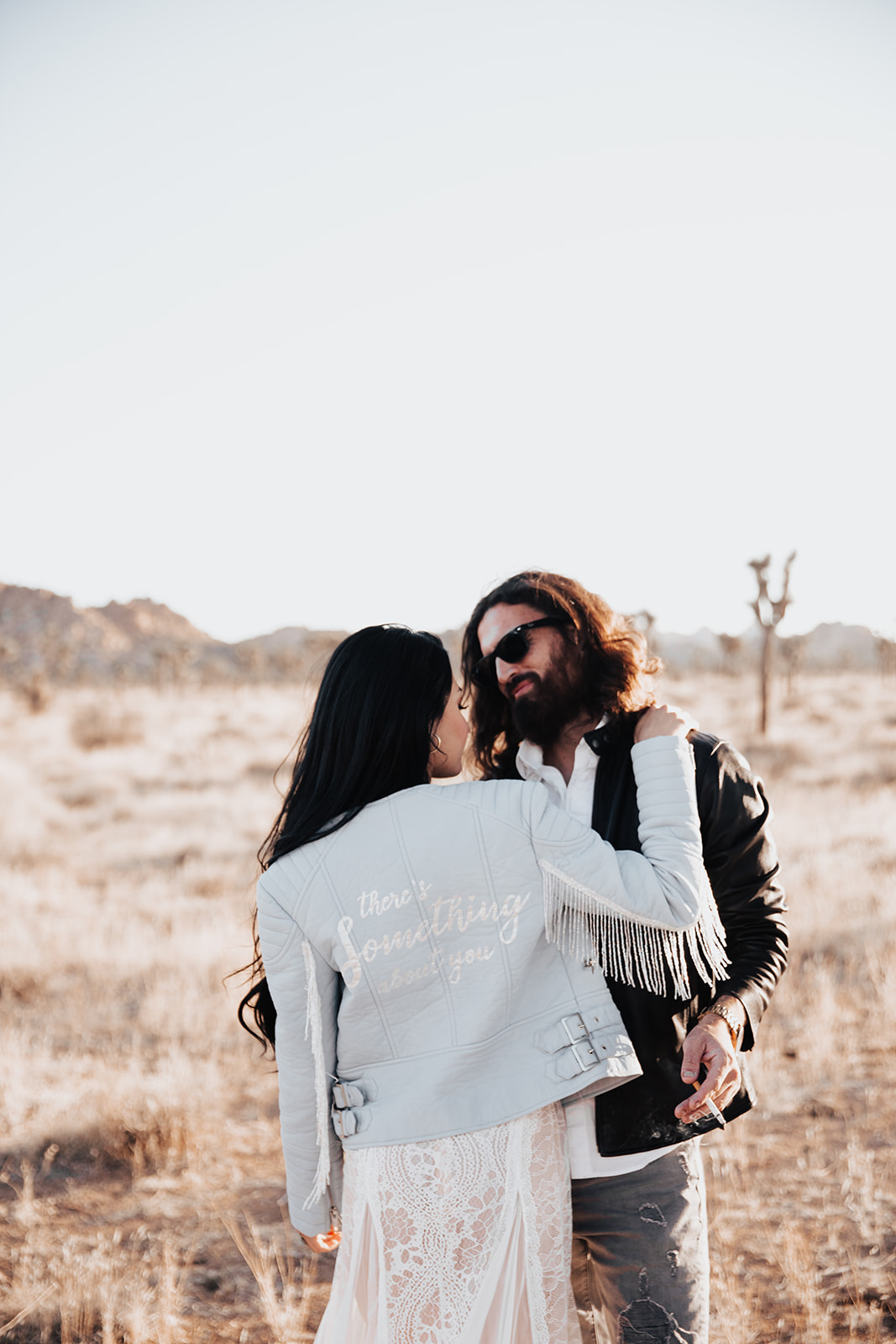 Long haired man with sunglasses and woman in white dress and light blue jacket standing in Joshua Tree for engagement