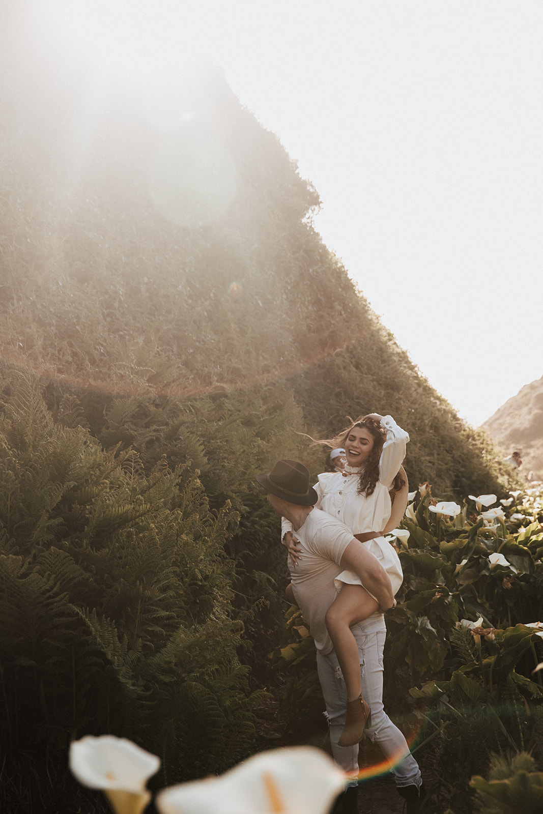 man gives fiancé piggyback walking through calla lily valley