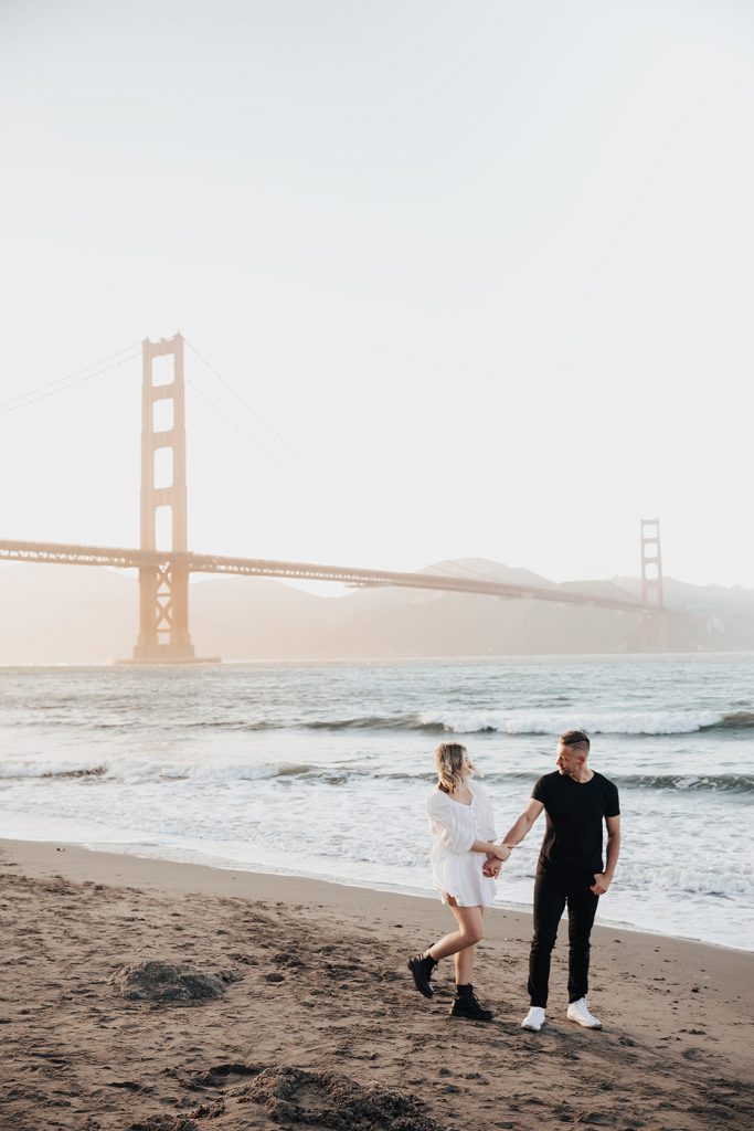 Couple walking the beach with golden gate bridge in background at sunset