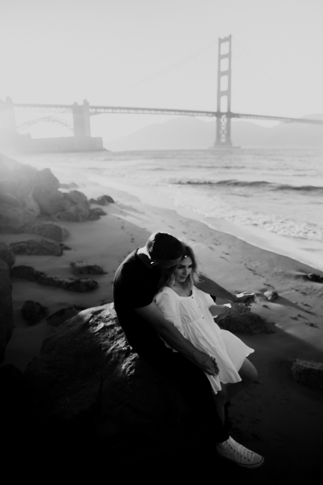 black and white photo of man in black shirt and woman in white dress at San Francisco Bay during dreamy golden gate engagement session