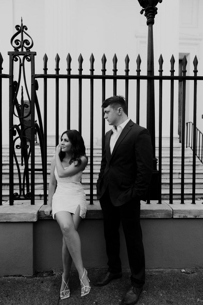 Couple in front of black fence in chic engagement session outfit. White dress and blue suite
