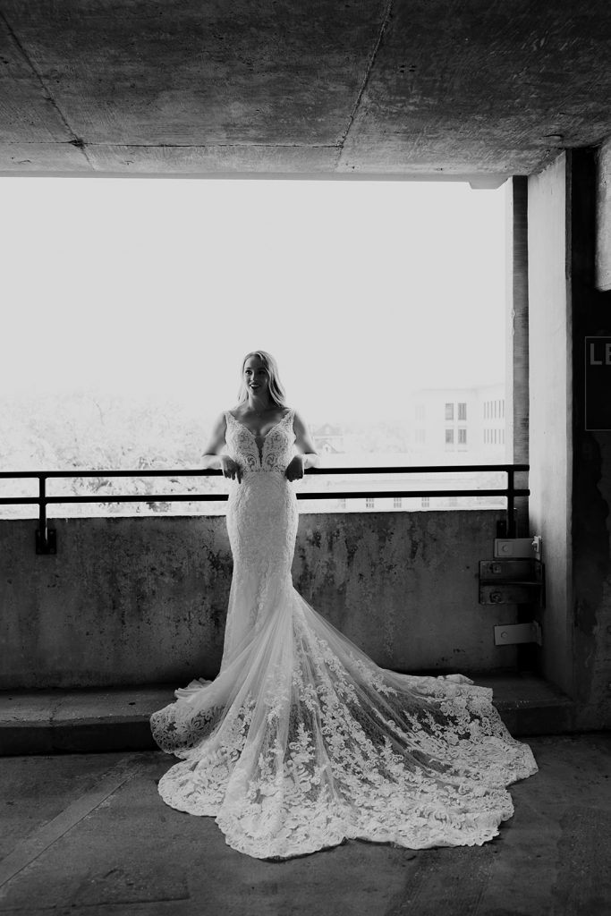 Bride leaning on railing in parking garage deck with beautiful dress. Black and white photograph. 