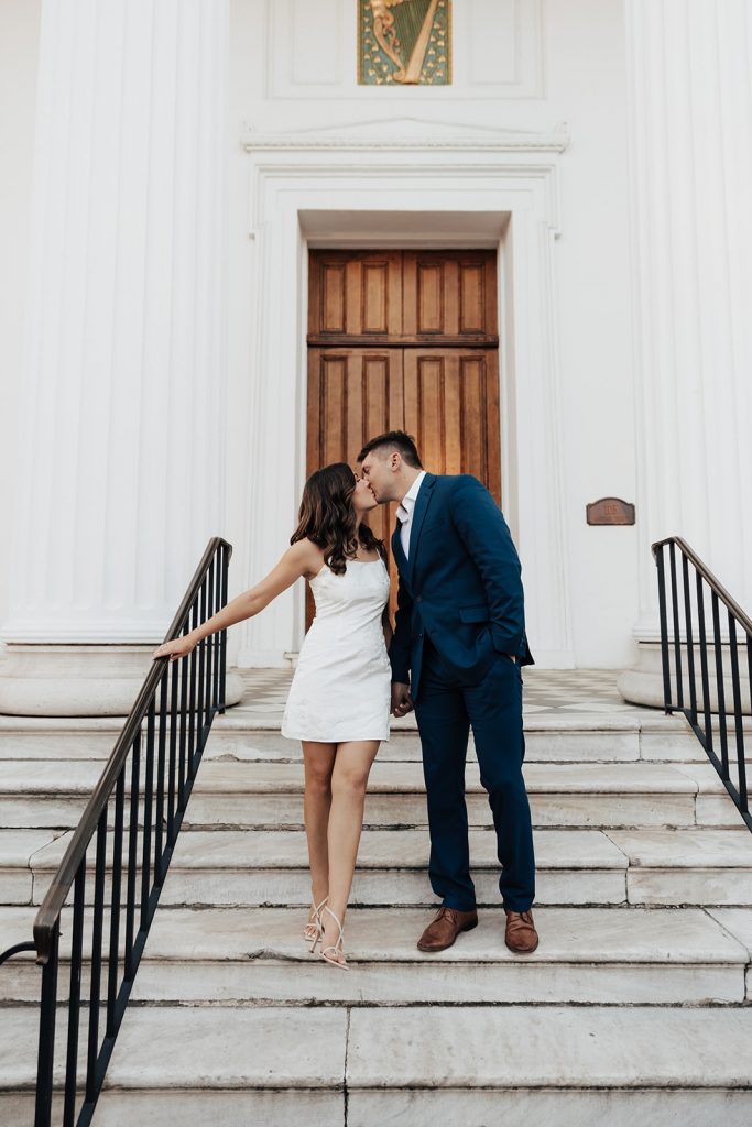 couple walking down stairs of Hibernian hall in chic white dress and suite during Charleston engagement session