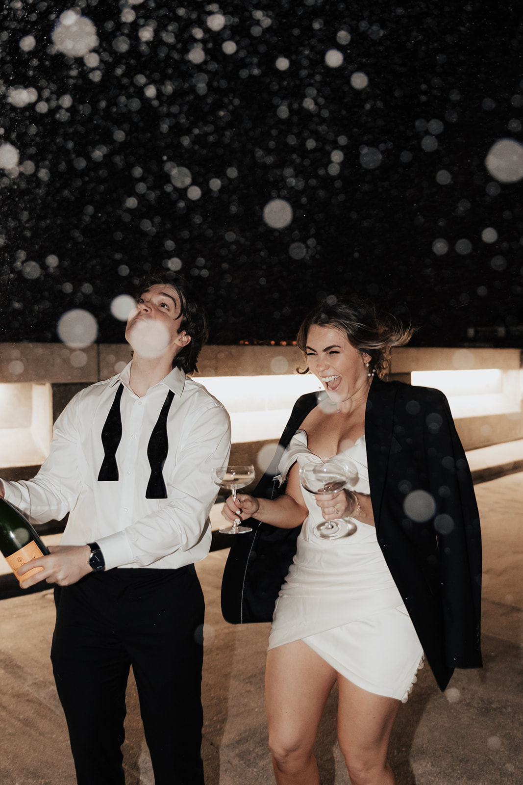 Couple sparying with champagne on top of a parking garage during Charleston engagament session. Woman hlding glasses and has jacket above her white dress. Man with white shirt holding the bottle.