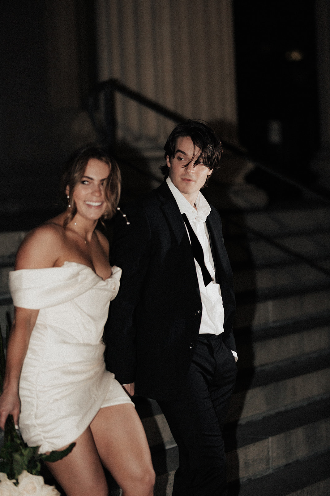 Couple walking. Woman in white dress holding on to bundle of roses. Man with bow tie untied and hair in his face. Both looking towards the camera for Charleston engagament session