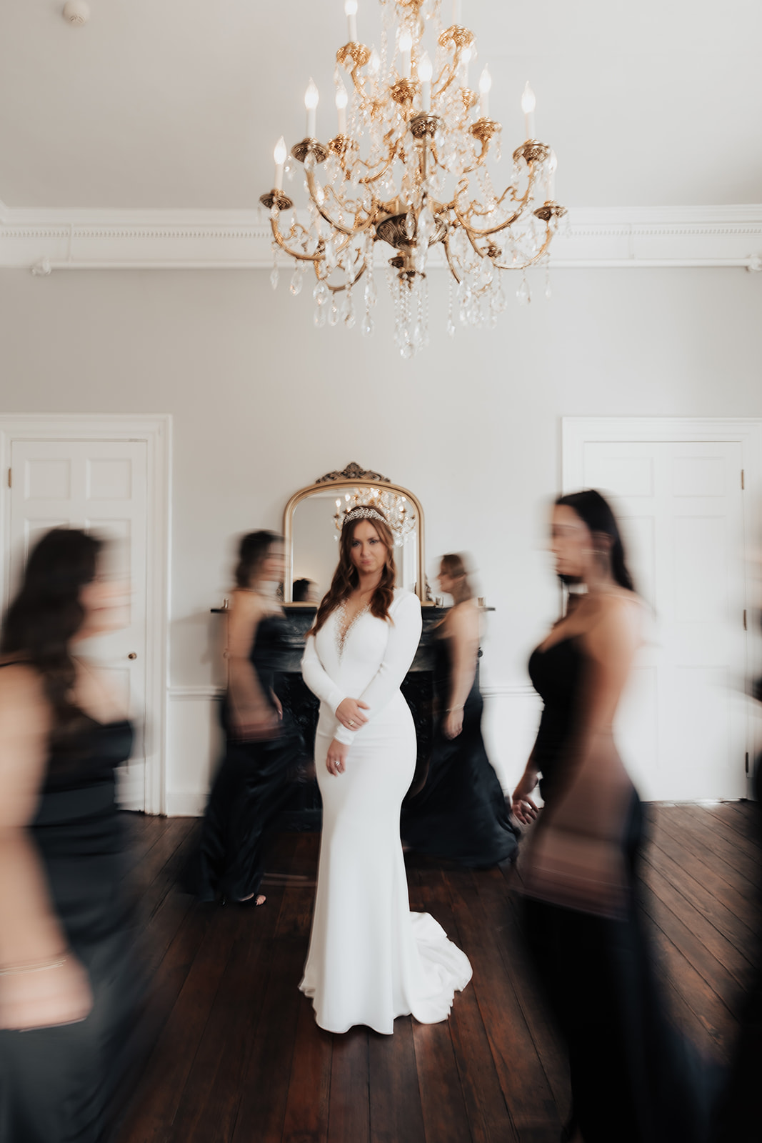 Artsy wedding photographer style captured bride in focus standing in middle of room surrounded by bridesmaids in black dresses all out of focus