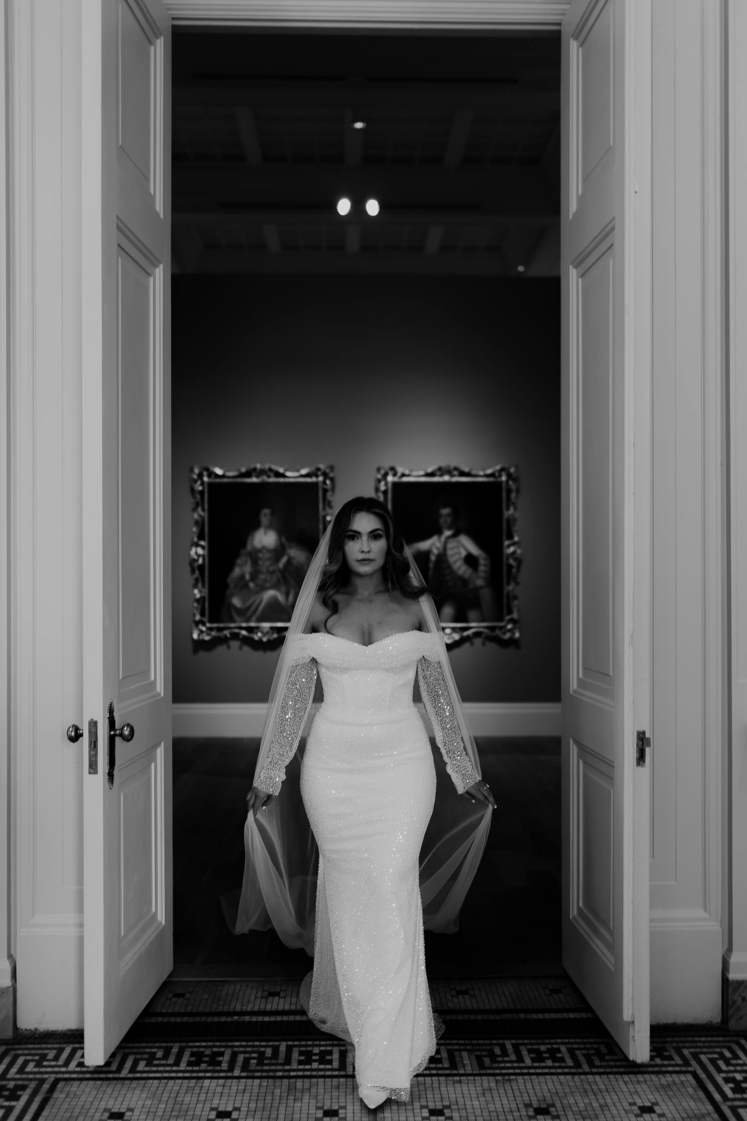 Black & white bridal photo of bride walking through door with paintings in background