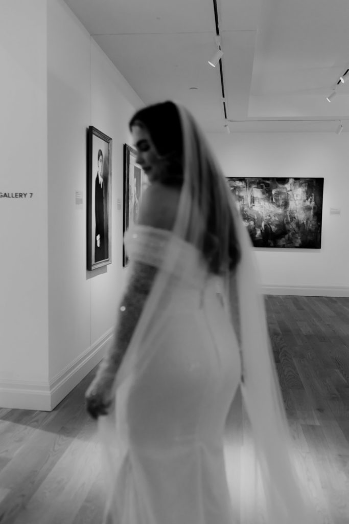 Black & white phot of bride out of focus in front of art and paintings