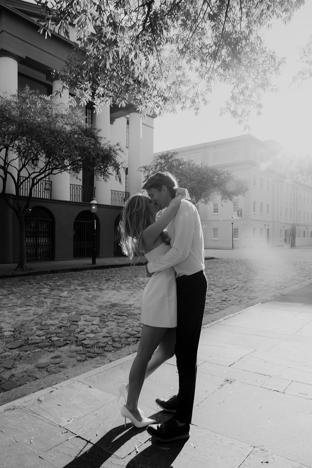 Black and white photo of couple standing on cobblestone street. Sun beams coming from behind