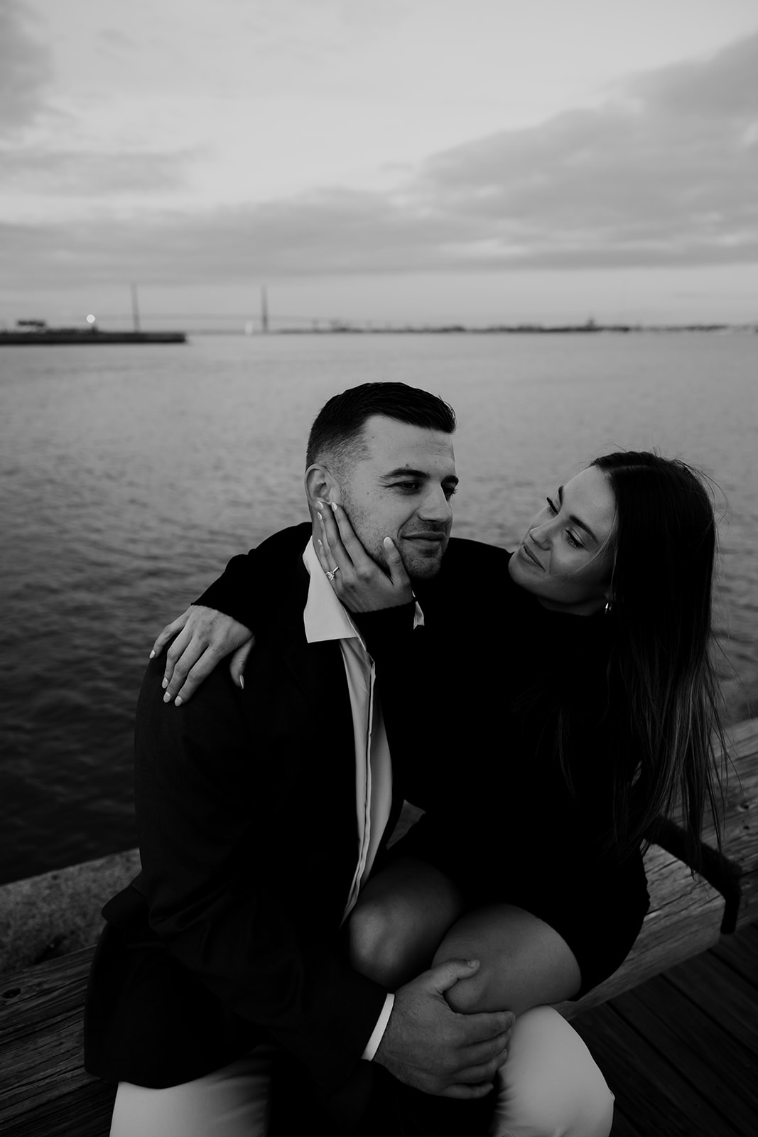 Black and white photo of couple sitting at public pier with bridge in background