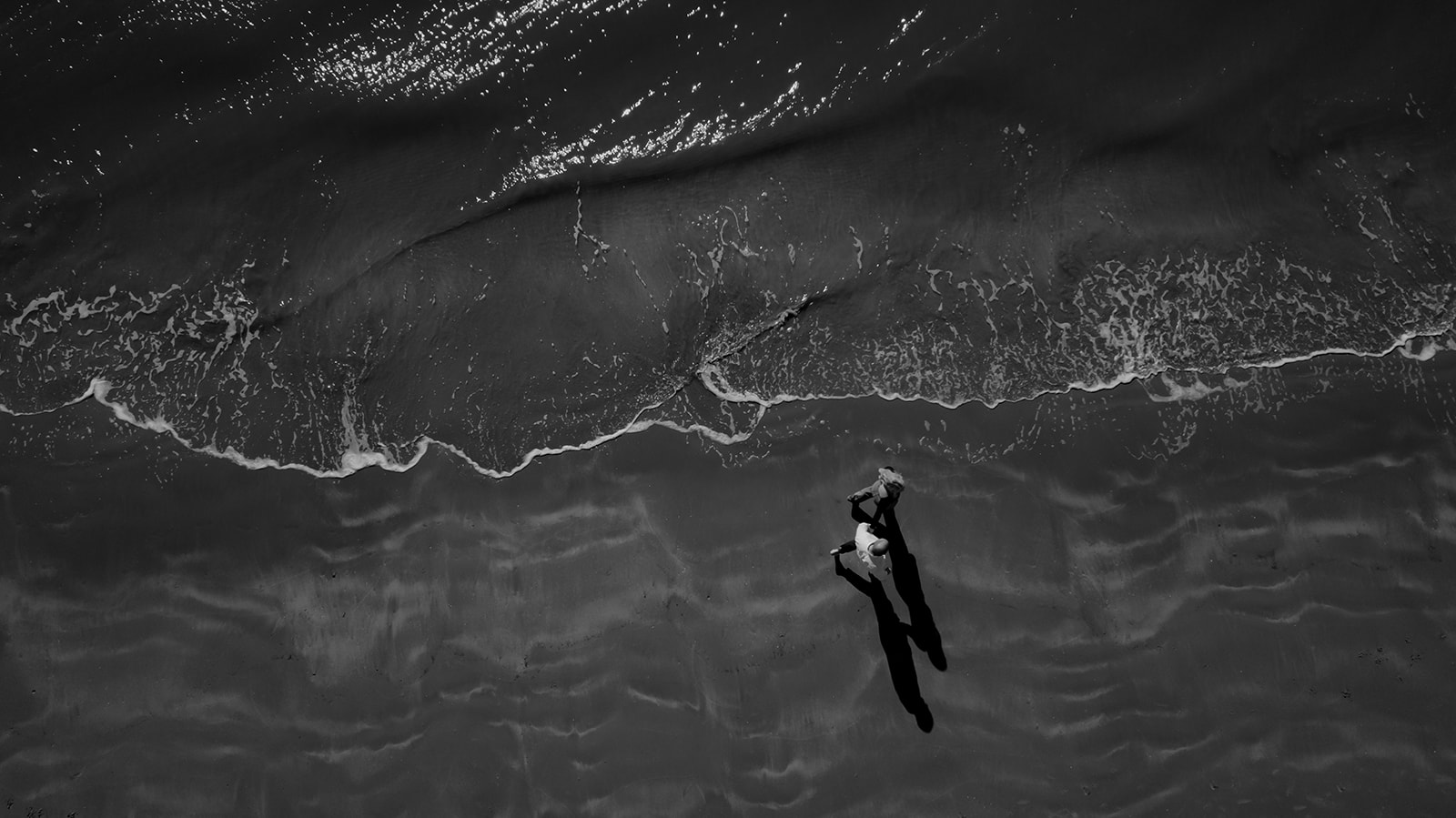 Seabrook Island Engagement photo session photographed from above. Waterline at the top of the photo and couple walking along it on beach.