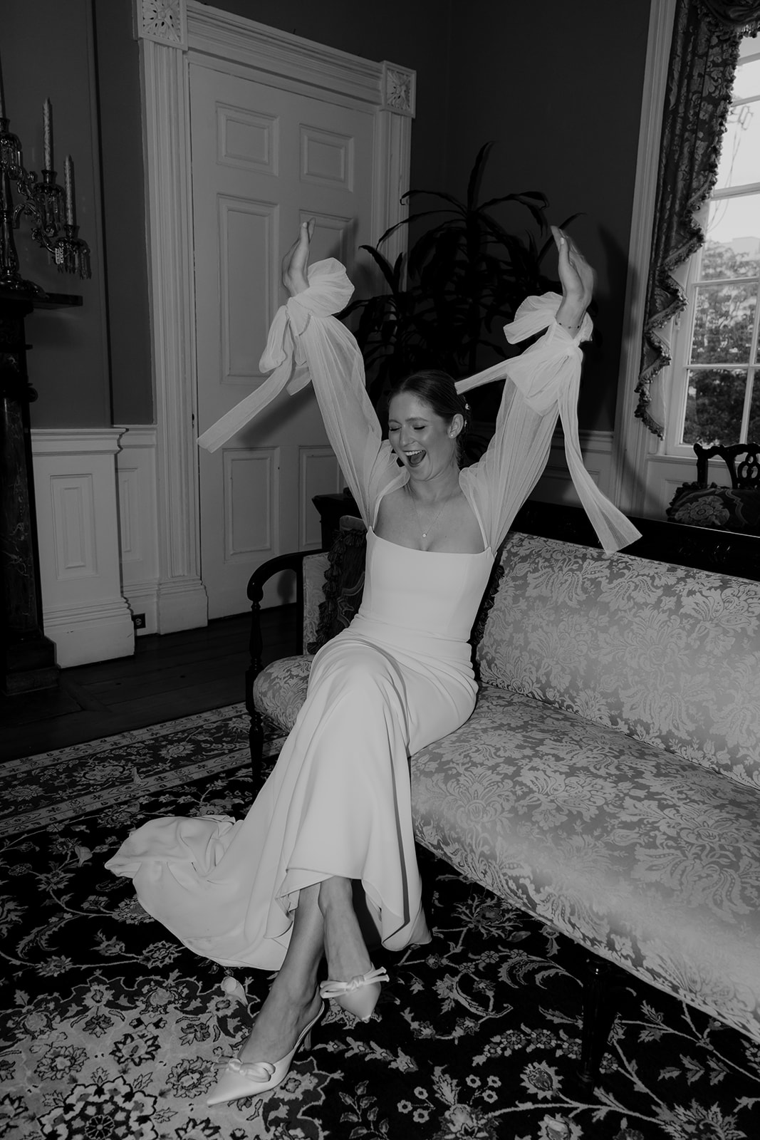 Bride sitting on french sofa and cheerfully raises both hands. Photographed in black and white during Wedding at Gov. Thomas Bennett House