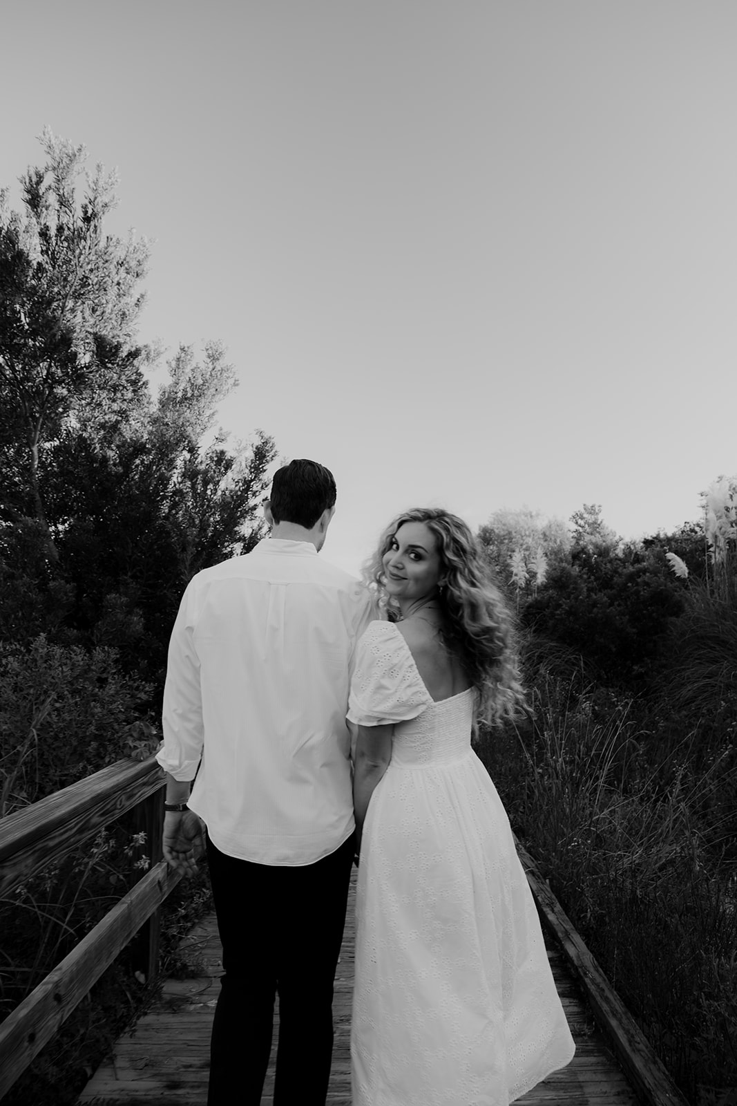 Black and white prortrait of couple walking towards beach on wooden boardwalk. Woman is looking back at camera