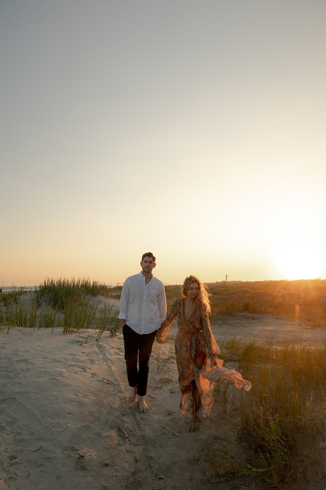 couple walking through dunes for Charleston Beach Engagement Photo with sun in back