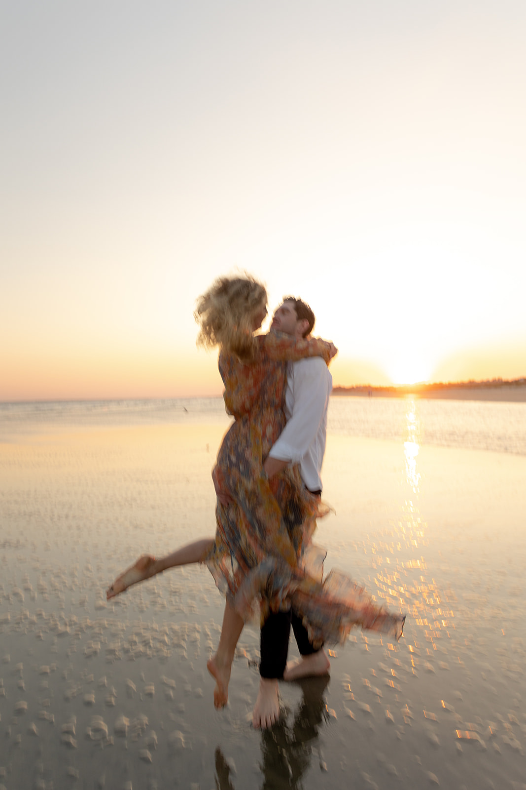 Charleston Beach Engagement Photo. Man picks up woman and twirls in low water with sunset in background