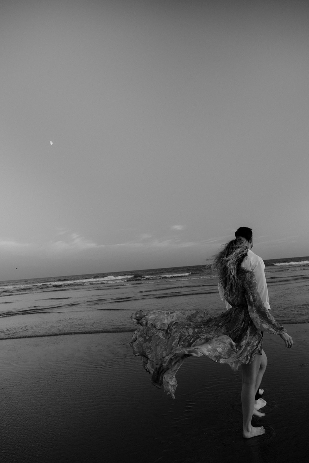Wind blows dress at Charleston Beach Engagement Photo in black and white. Couple walking close to water