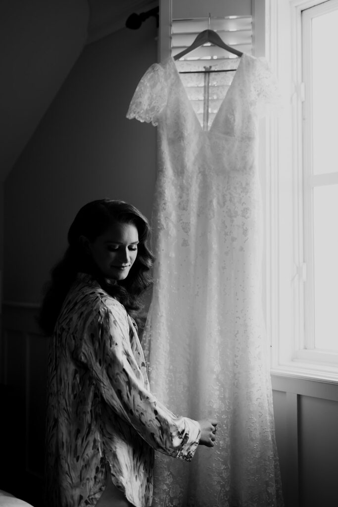 Black and white photo of bride looking at dress hung at window
