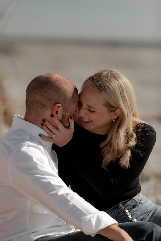 Couple sitting in sand. Woman holds man's head and laughs during Seabrook Island Beach Engagement