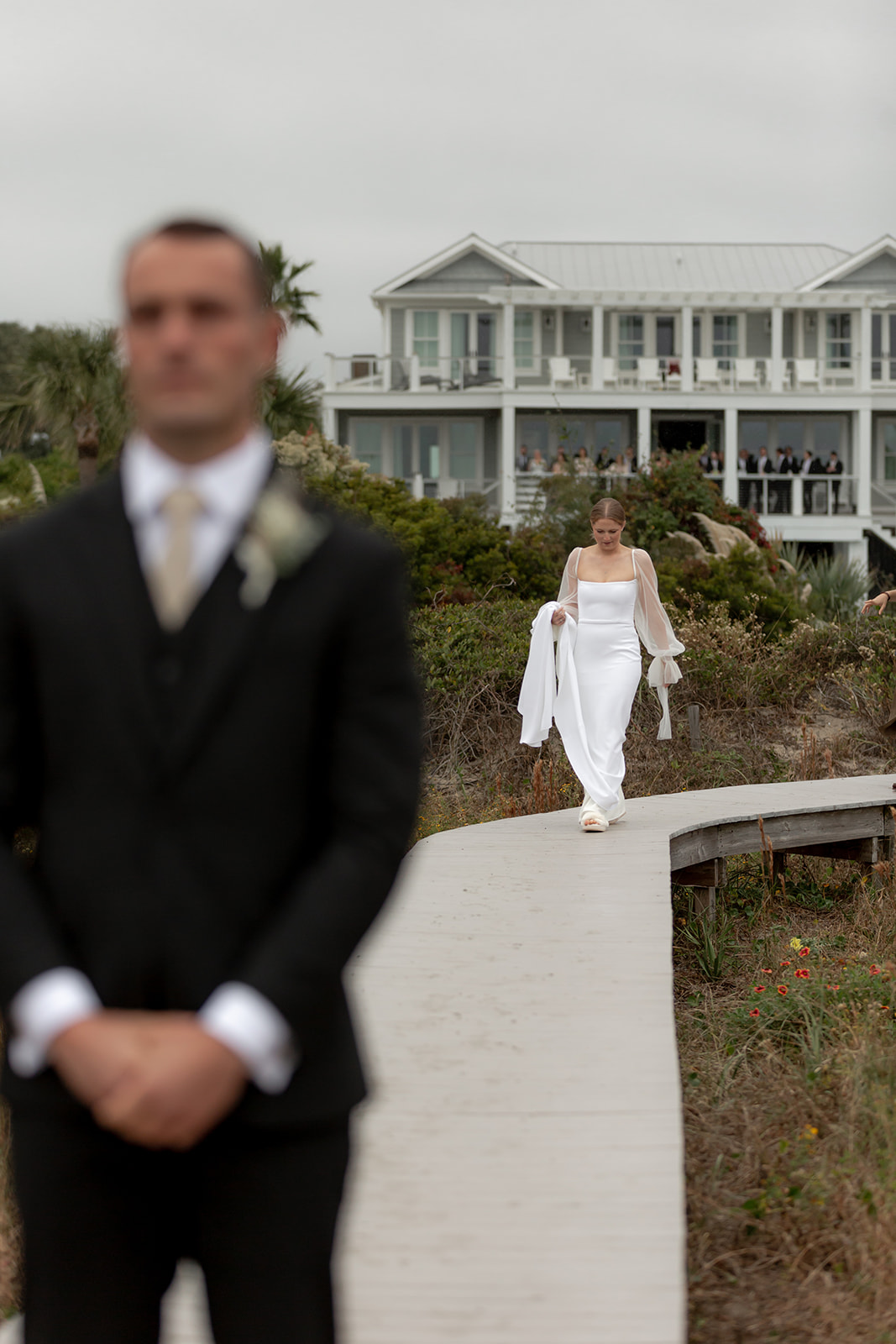 Before the first look. Bride in the background and in focus walking along wooden boardwalk towards groom. He stands in the foreground and out of focus with his back turned to the bride.