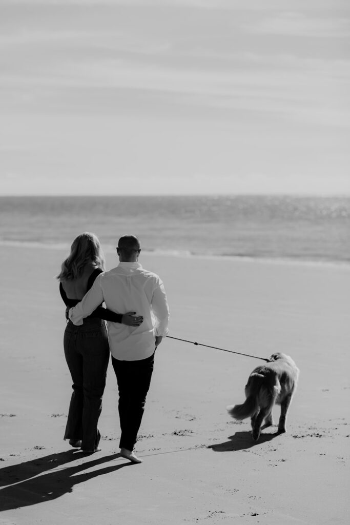 Couple with arms wraped around each other walking towards water. Dog on leash walking in front.