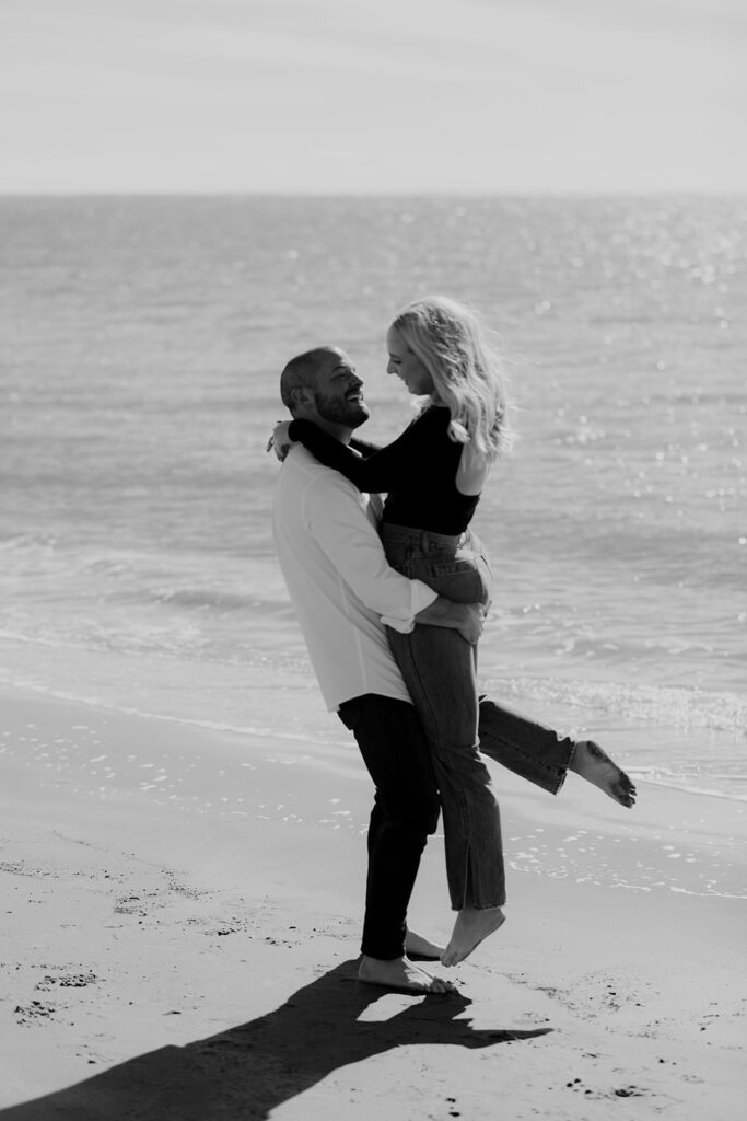 Man holding up woman at the beach. Ocean in the background. Black & white photo