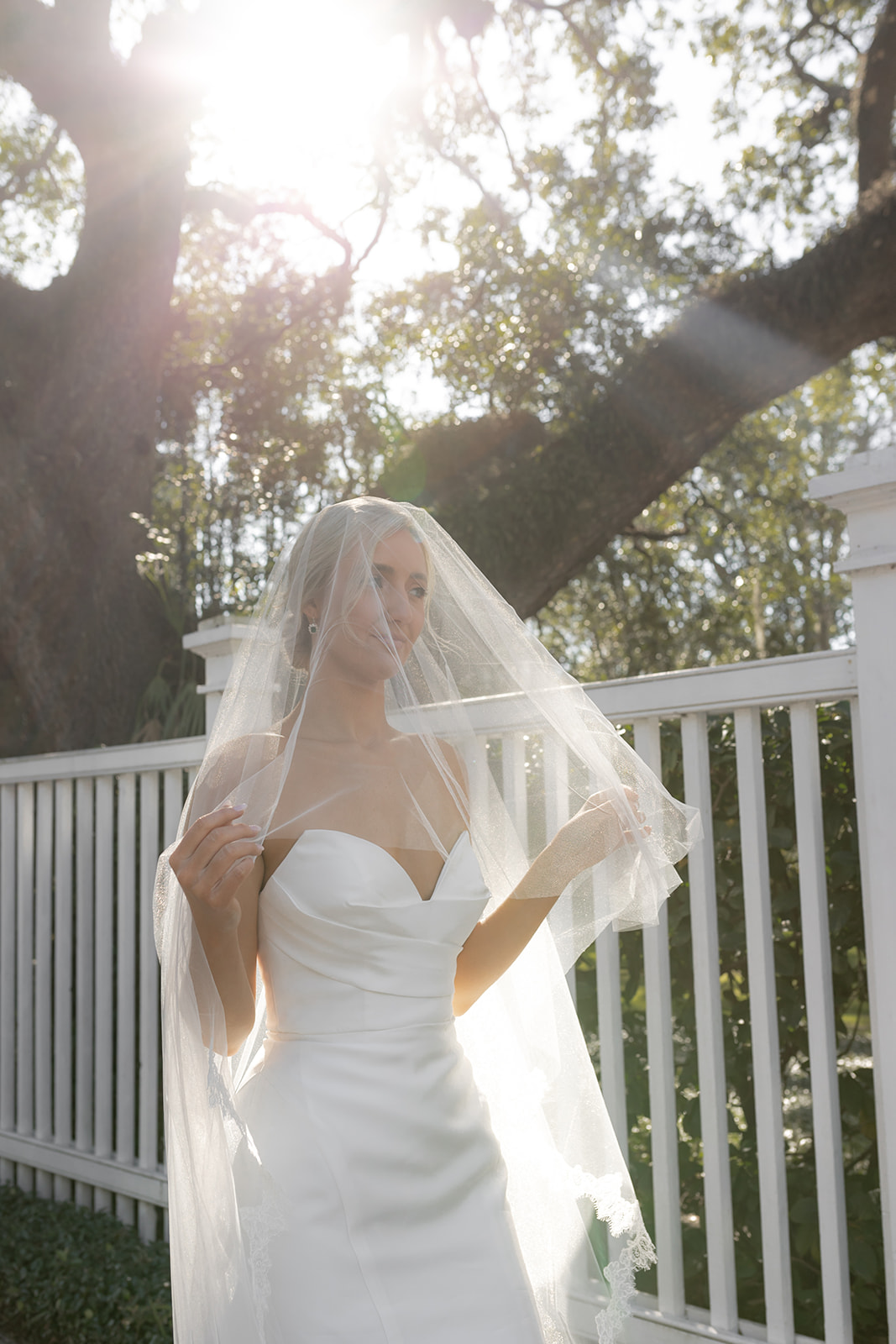 Bride walking outside. White fence and oak trees in background sunlight hitting wedding veil. Perfect situation to show wedding dress in Charleston