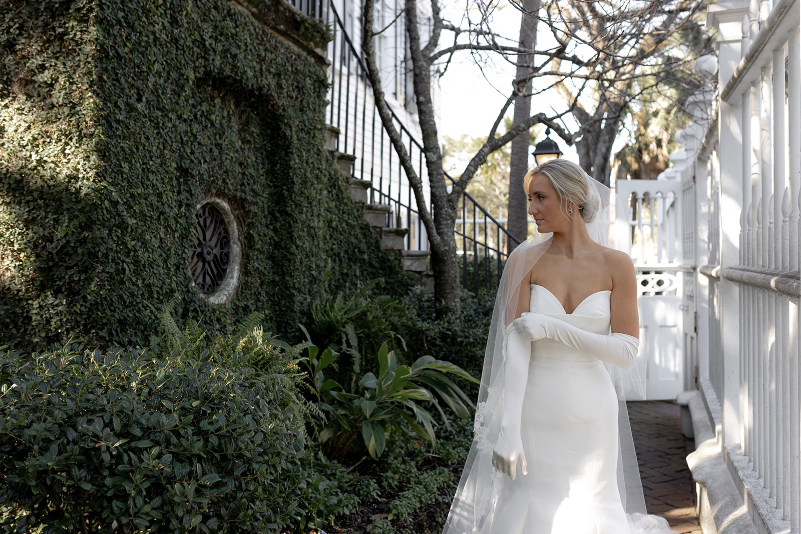 Bride adjusting long-sleeves gloves of wedding dress. Standing next to ivy grown wall with ornamental window at Gov. Thomas Bennett House in Charleston.