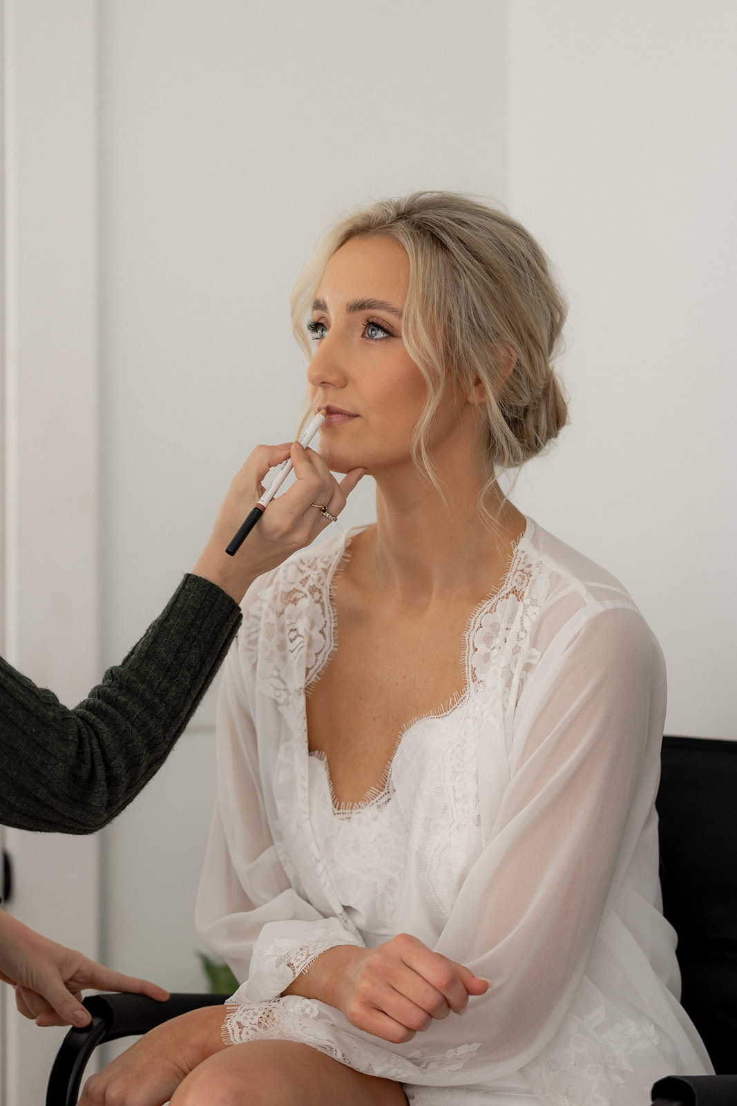 Bride getting ready for wedding. Sitting on chair in white robe while lipstik is applied