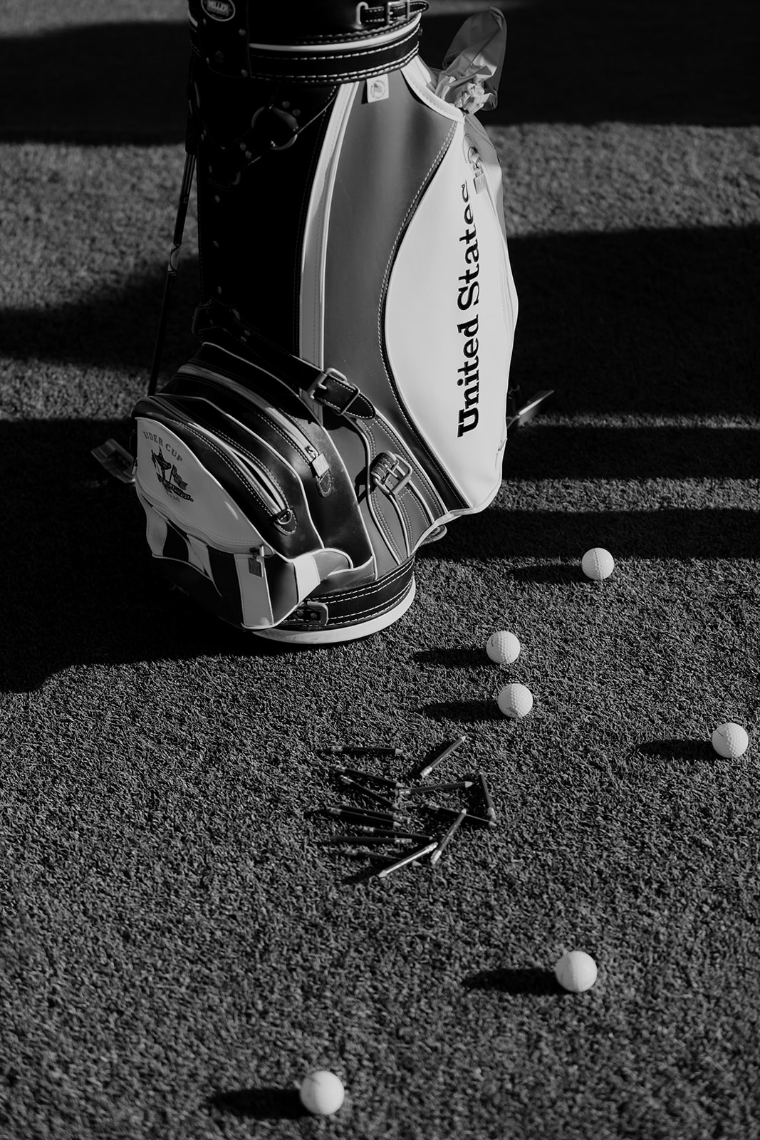 Black and white photo of golf bag with golf balls and tees on the floor.