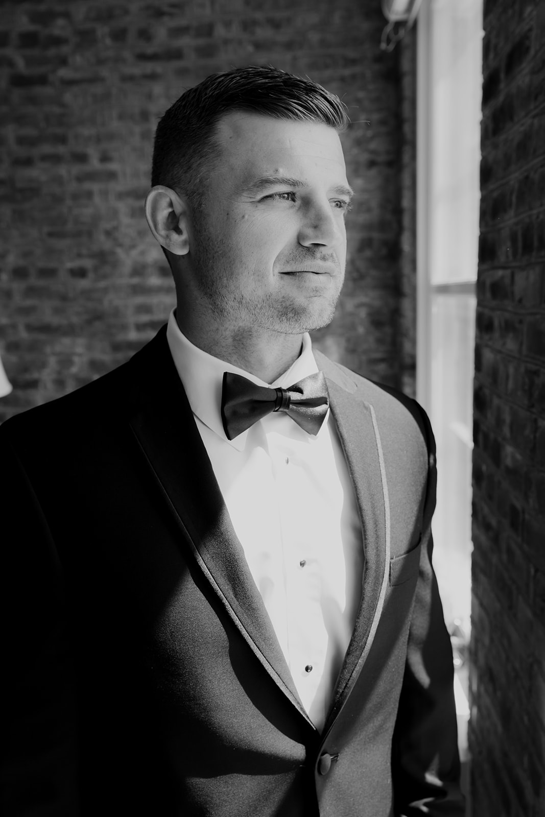 Black and white photo of groom in black tuxedo with bow tie looking out of the window.