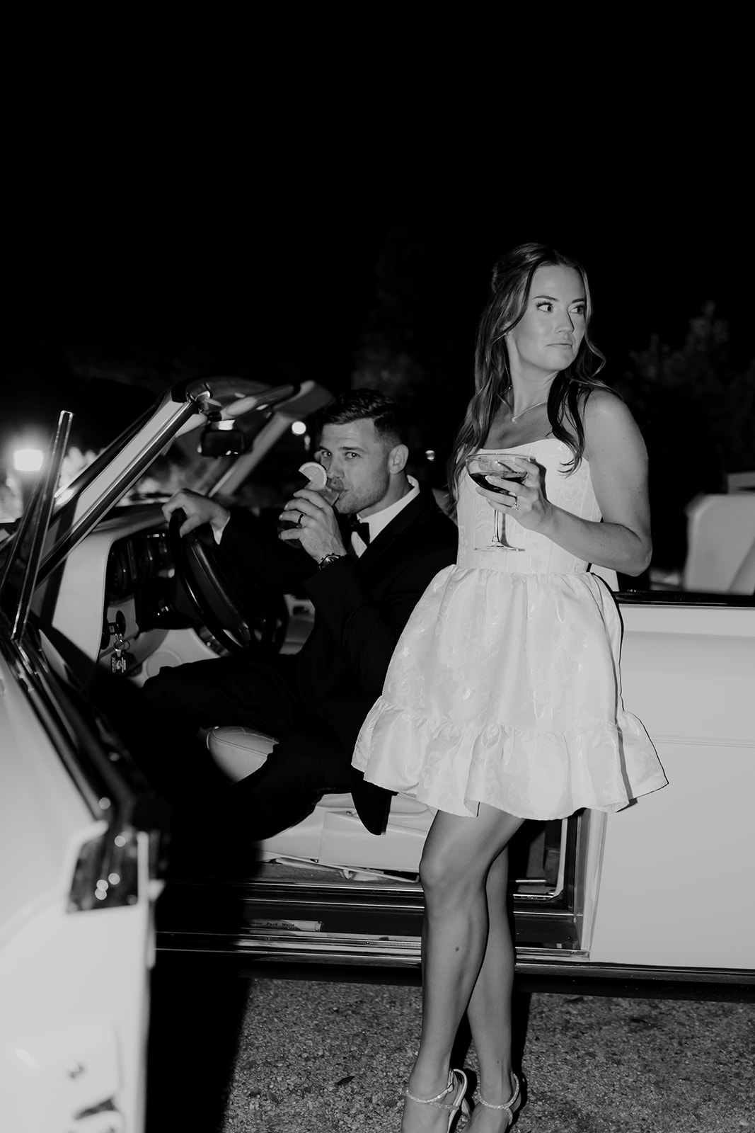 Black and white photo of couple and their weddign get-away-car. Groom sitting inside and bride standing at door.