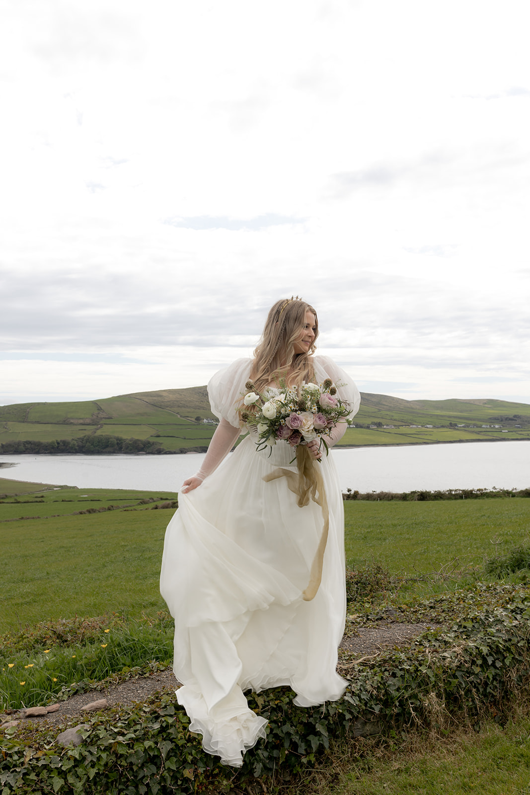Bride posing on small wall for irish elopement photos with water and hills in the background