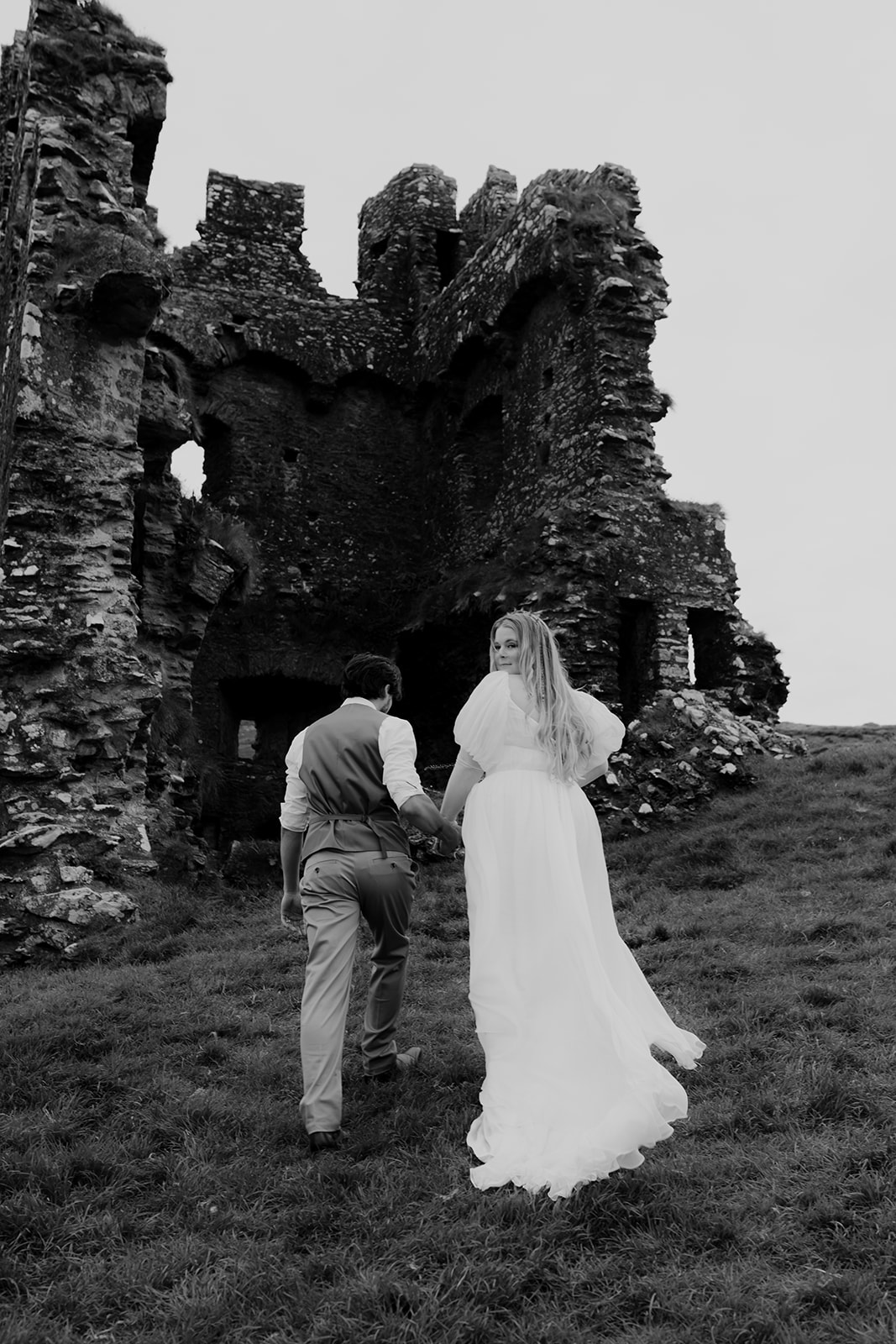 Bride and groom walking towards castle ruins. Bride looking back at camera.