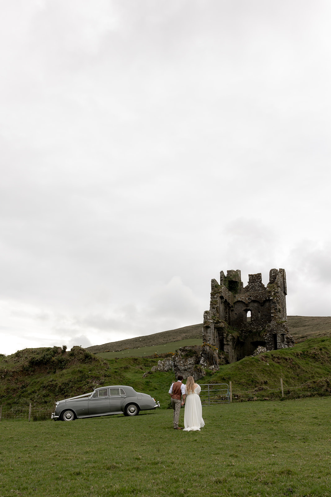 Bride and groom walking towards castle ruins. Rolls Royce parked next to them.