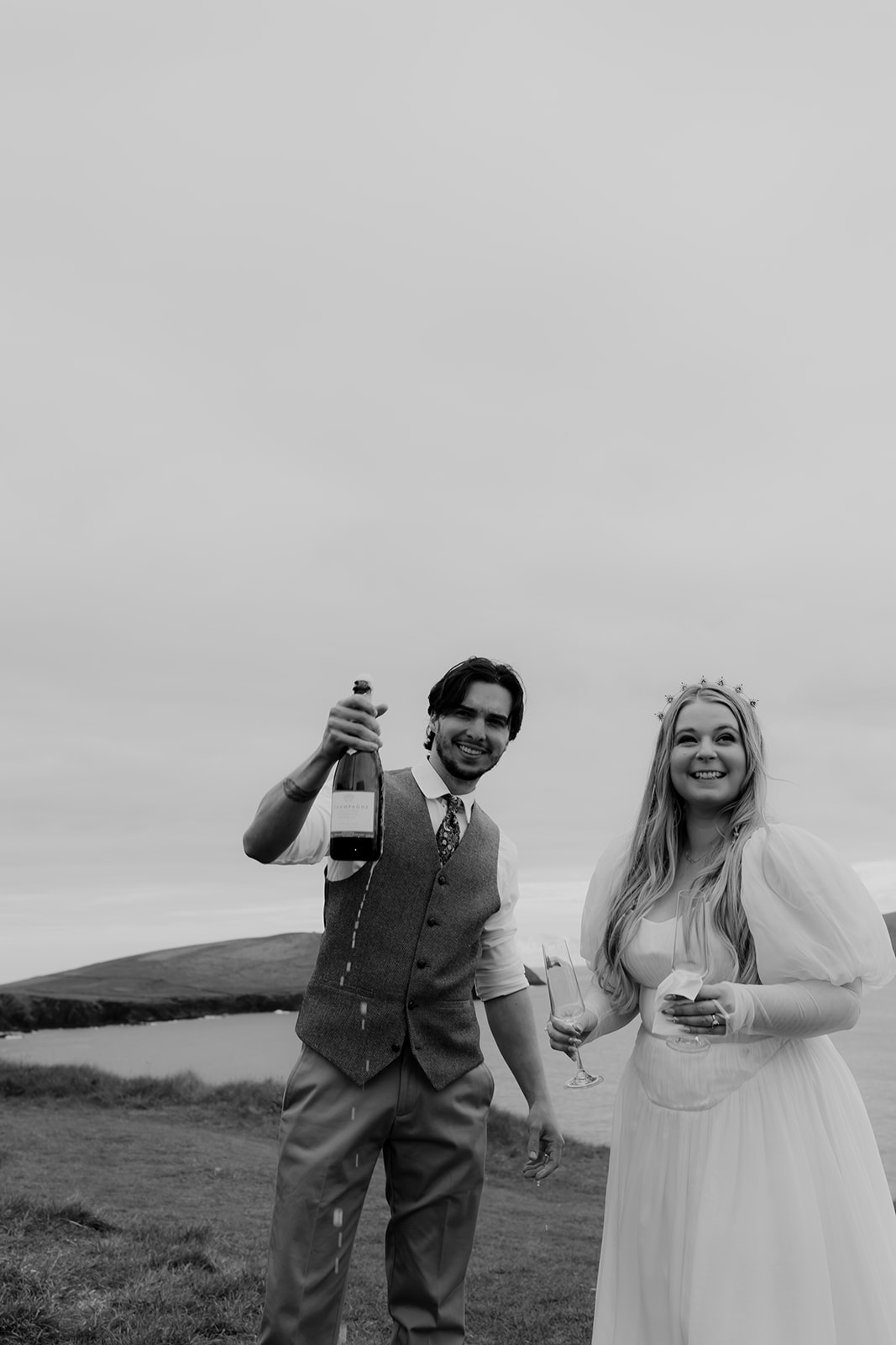 Groom opened bottle of champagne. Bride smiles and holds glasses to drink on Ireland elopement