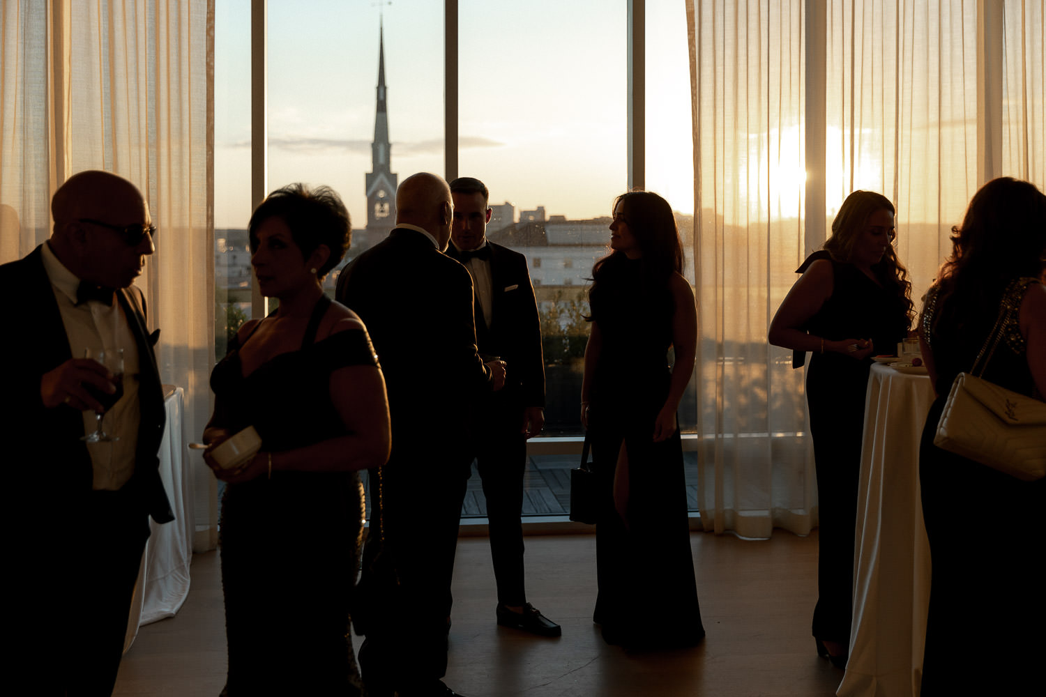 Guests socializing at the Rivers Room at a Dewberry wedding. Sun is setting in the background creating silhouettes of people