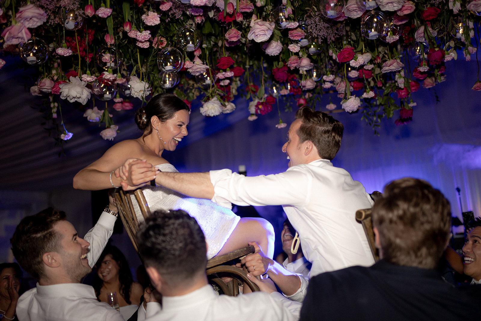 Bride and groom celebrating the hora or chair lift celebration under a hangig flower arrangement with red and pink flowers at Lowndes Grove wedding