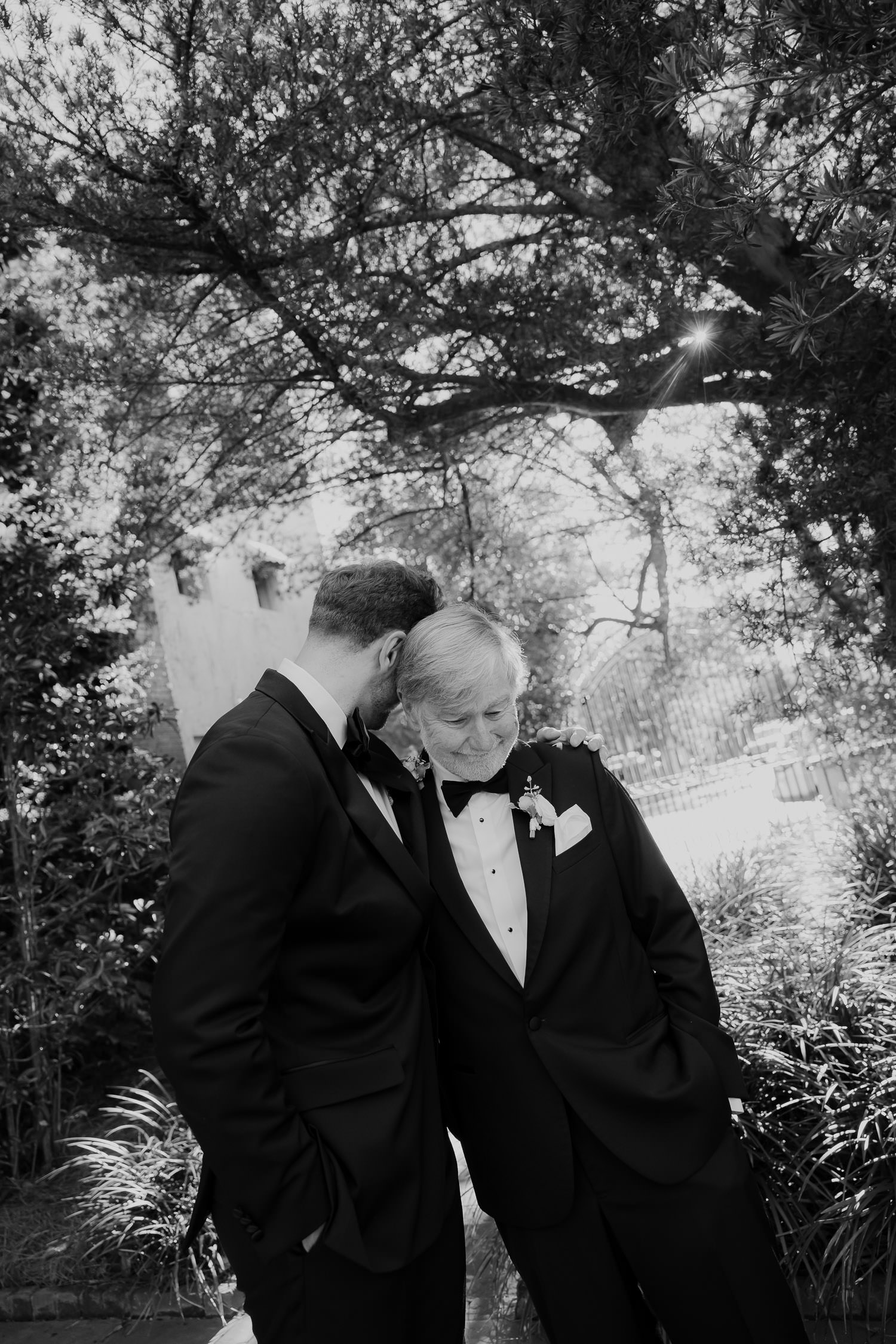 Black and white photo of intimta emoment between groom and his father standing underneath live oak.