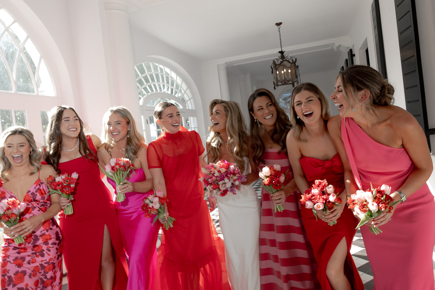 A Hot Pink and Red Wedding Extravaganza. Bride and bridesmaids walking towards camera. The white dress is surrounded by gowns in all shades of red and pink