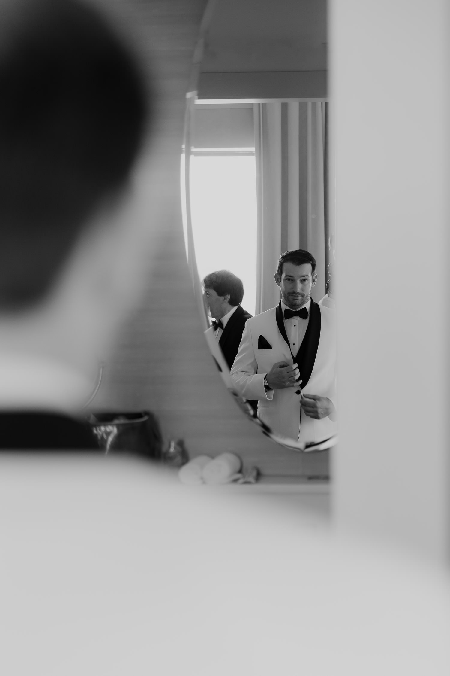 Groom getting ready at the Dewberry Hotel for a Winter Wedding in Charleston. Looking at mirror in white tuxedo jacket.