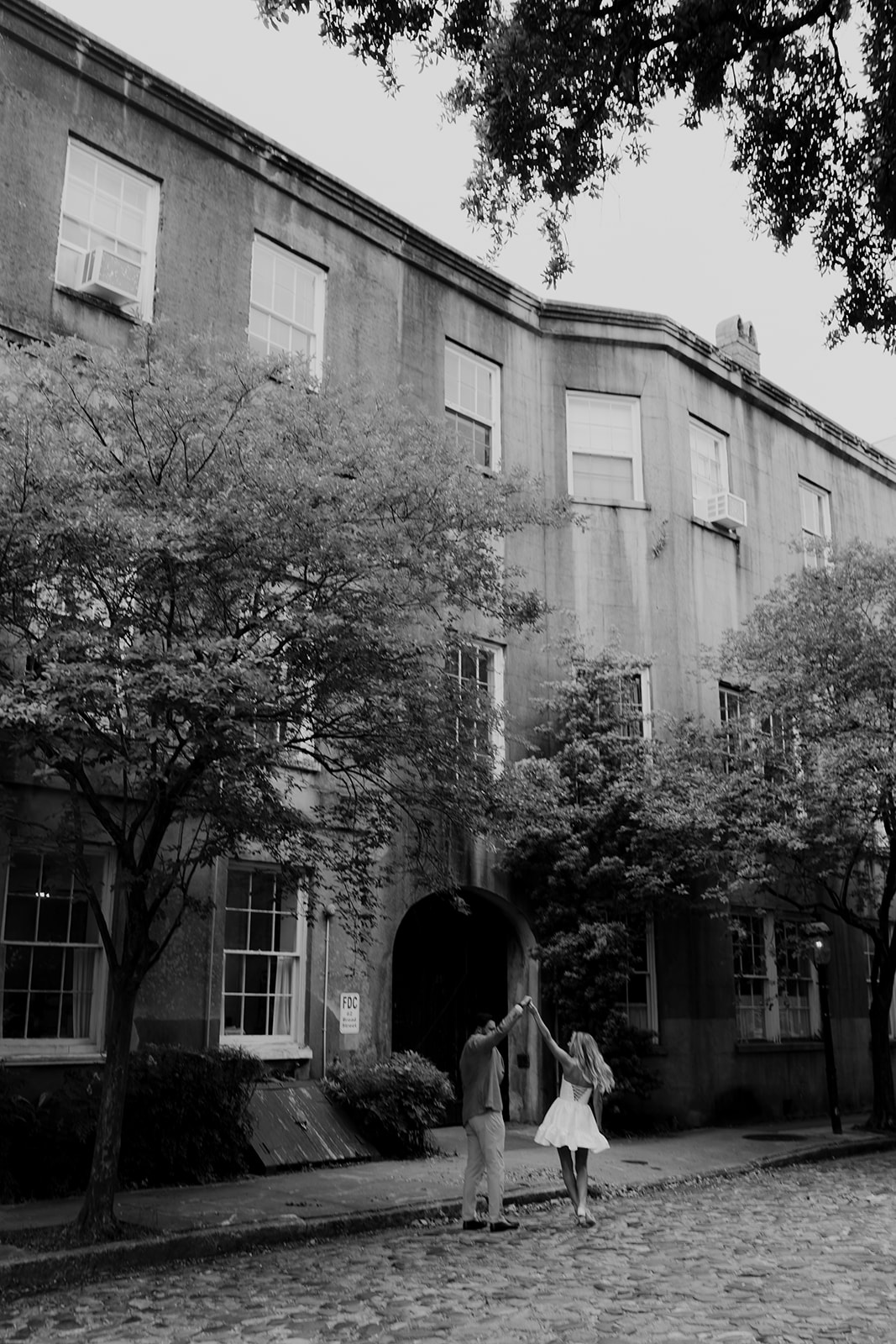 Black and white photo showing man twirling a woman in white dress on the CHarleston cobblestone taken during Downtown Engagement photo session