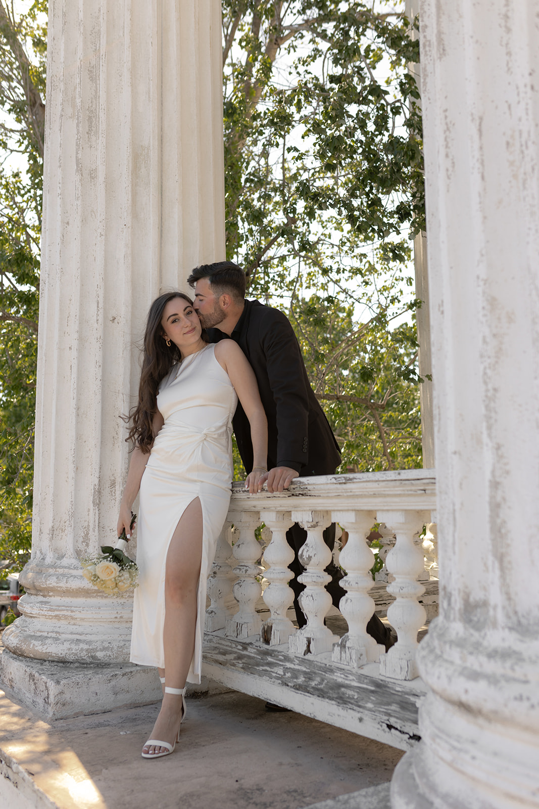 engaged couple in black and white engagement attire, standing between greek style columns. He gives her a kiss on the temple. 